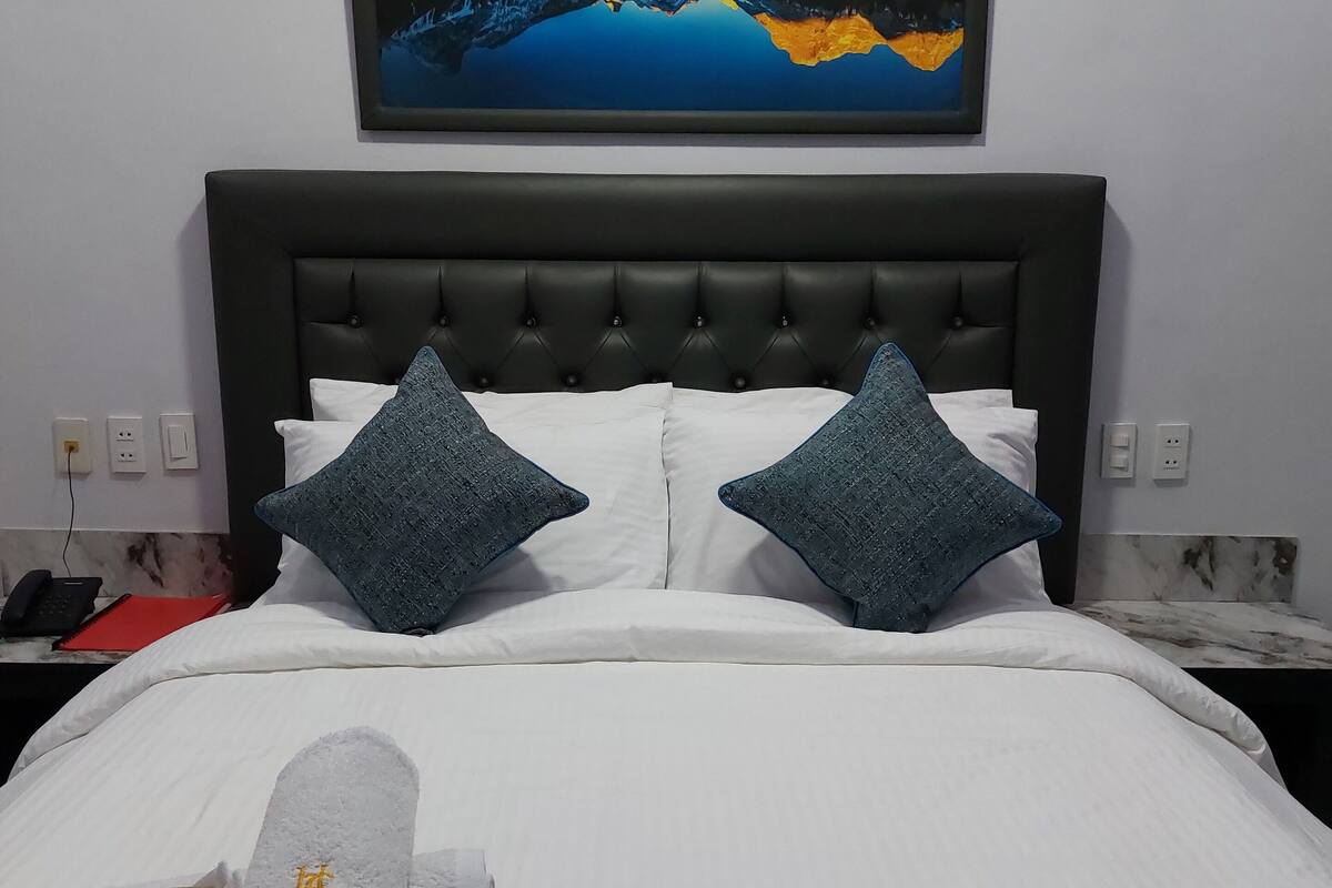 A neatly made bed with white linens and two blue pillows, a framed landscape painting above the bed, and a pair of rolled towels on the bed.
