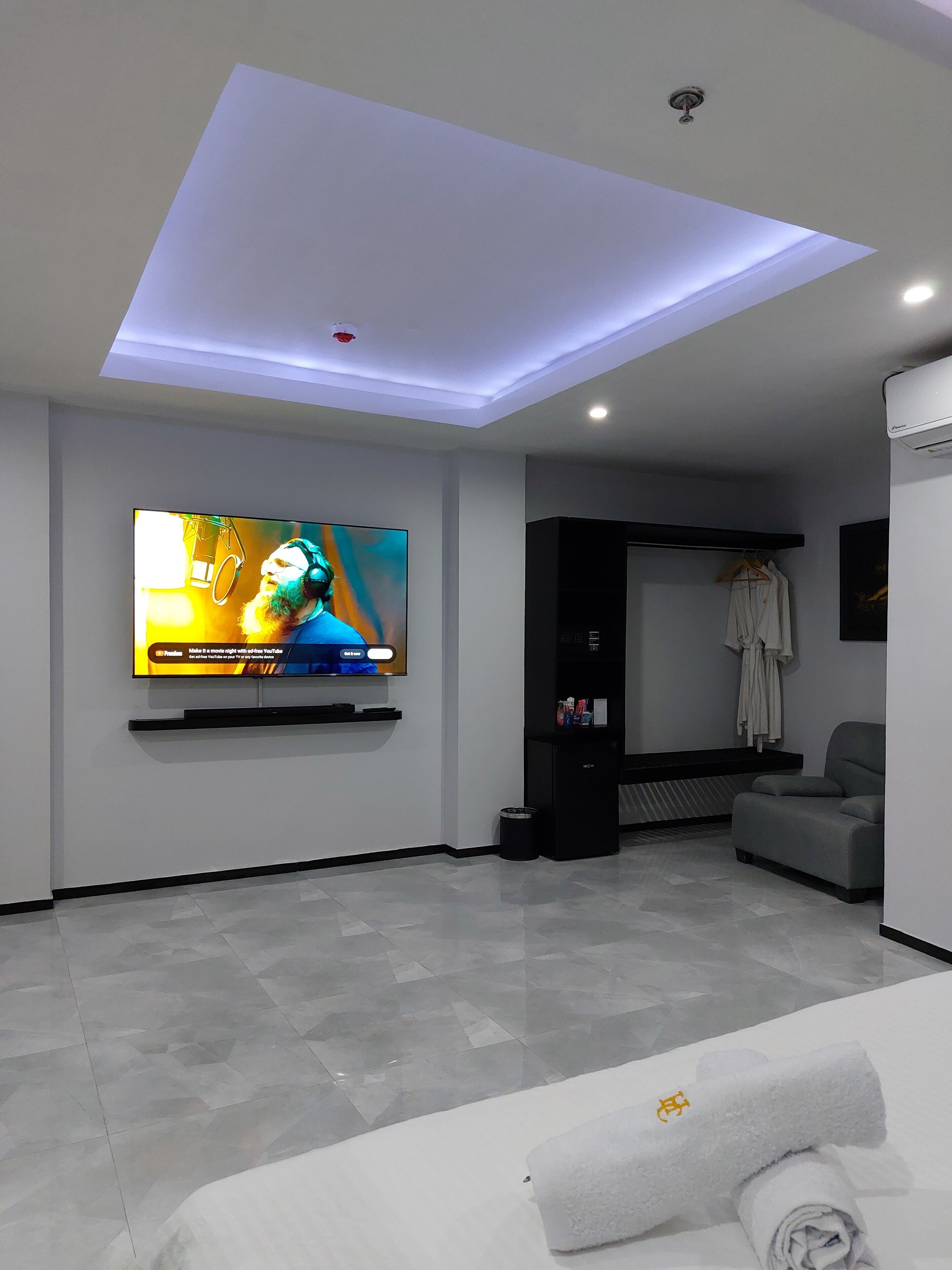 A modern hotel room with a flat-screen TV mounted on the wall, a ceiling light, a seating area with a grey sofa, and a small black cabinet.