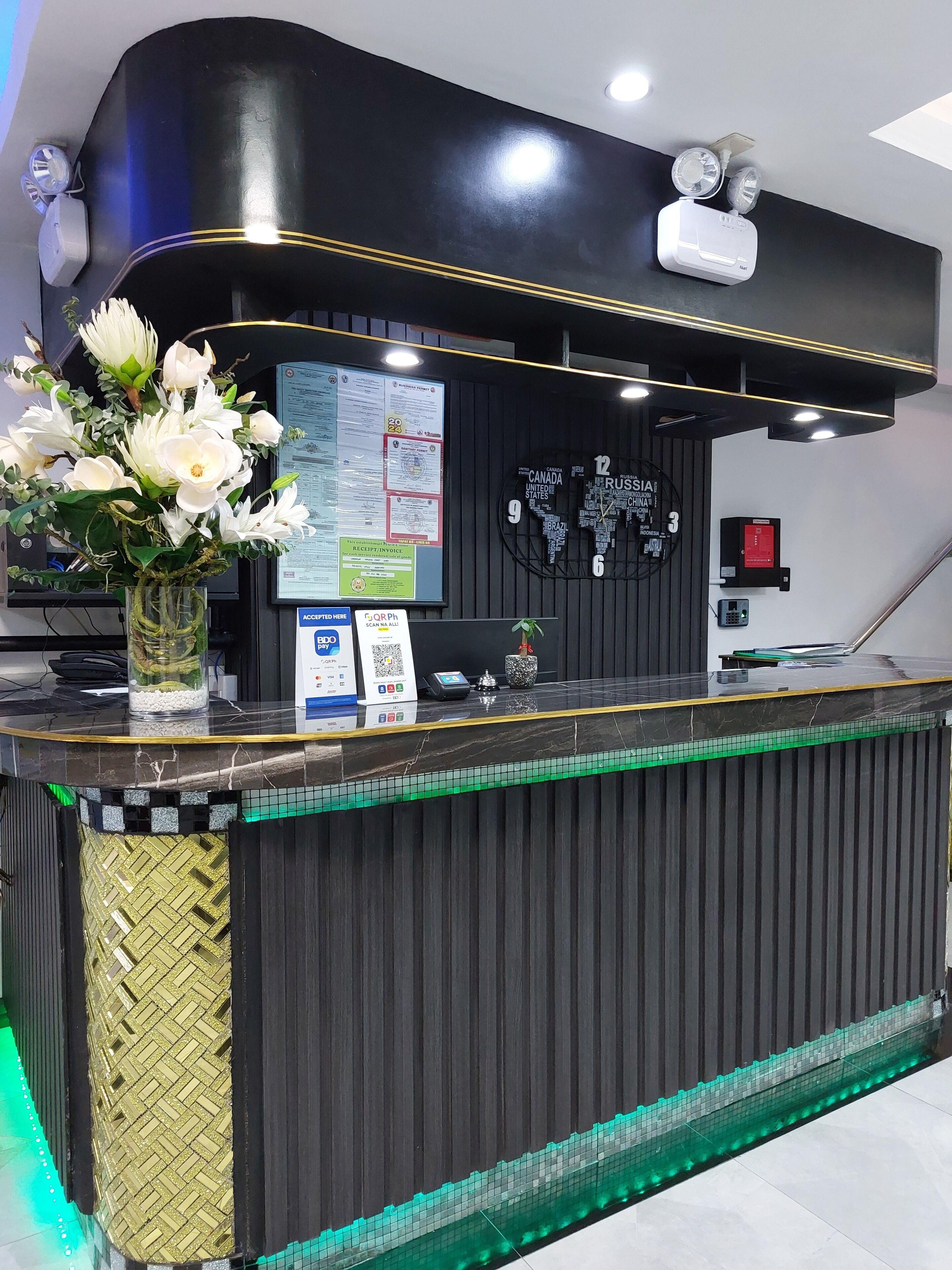 A reception desk with a black and gold design, a vase of white flowers, a digital screen, and a wall with country names.