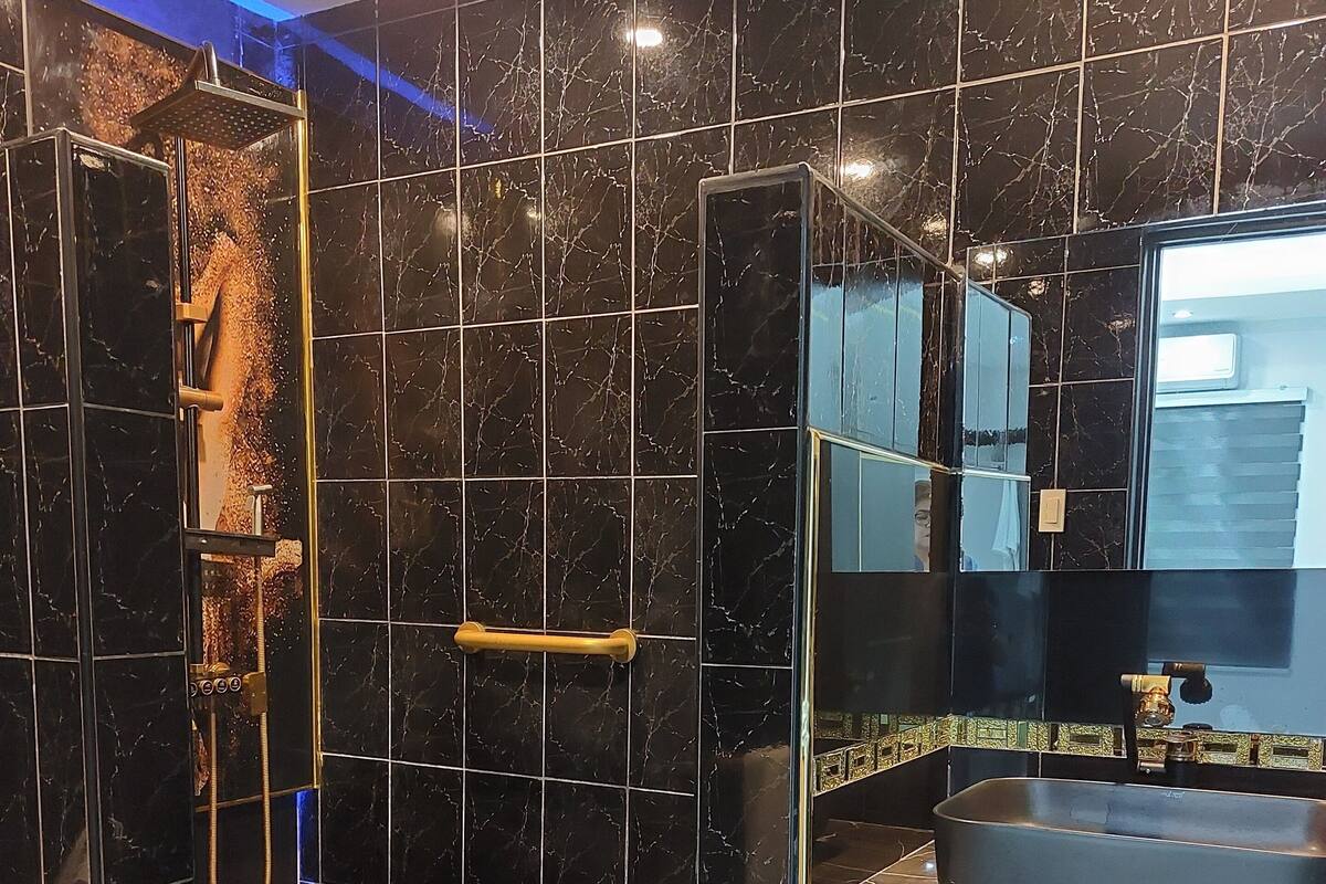 A modern bathroom with black marble tiles, a shower with a gold handle, and a sink with a mirror above it.