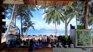 A beachside stall with various items for sale, including pots, cups, and a painting, under a thatched roof with palm trees and the ocean in the background. - Three ladies (Ko Yao)