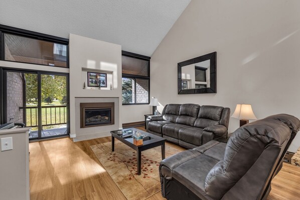 A modern living room with a fireplace, leather sofas, and large windows offering a view of the outdoors. - Park City Golf Course Condo: Pool & Hot Tub Access (Park City)