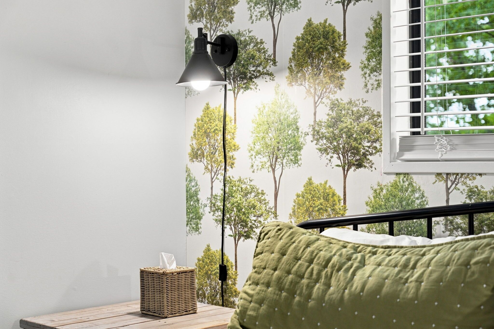 Une chambre à coucher avec un lit, une table de chevet avec une lampe, un panier et une fenêtre avec des stores.