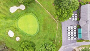 Vista aérea de un campo de golf con un gran green, búnkeres de arena y un estacionamiento con autos blancos y un edificio de techo azul.