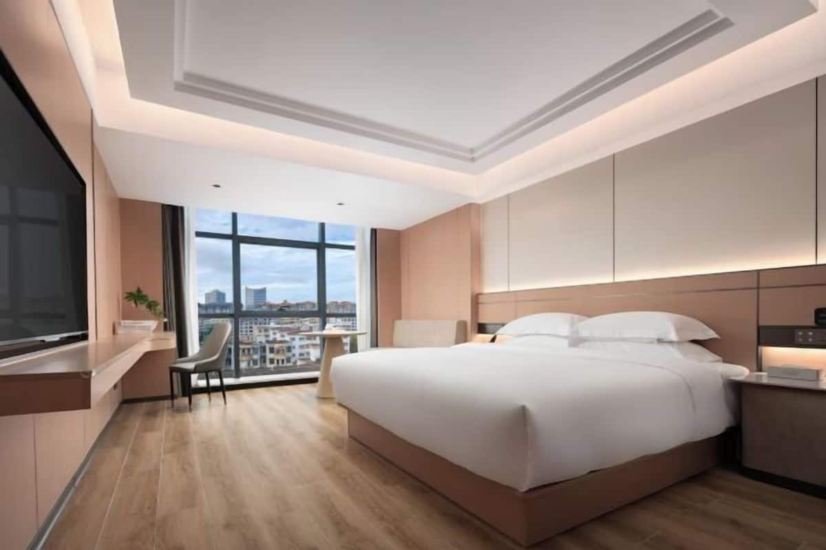A modern hotel room with a large bed, a desk, a chair, and a view of the city through large windows.