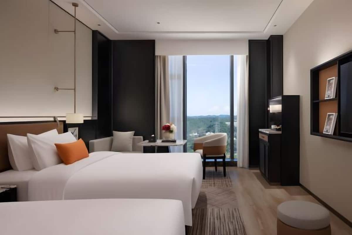A hotel room with two beds, a desk, a chair, a TV, and a large window with a city view.