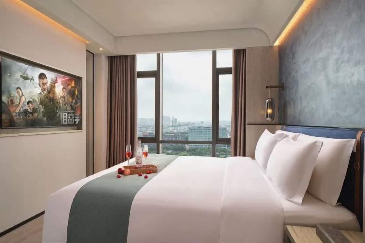 A hotel room with a large bed, a city view, a painting on the wall, and a nightstand with a bottle and glasses.