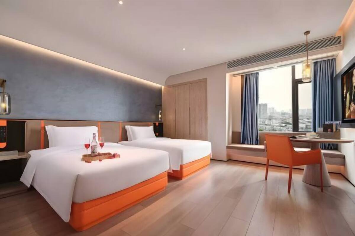 A modern hotel room with two beds, a dining area, and a large window with curtains.