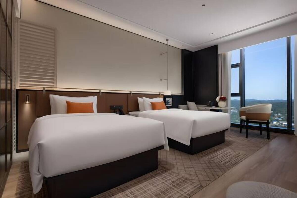 A modern hotel room with two beds, a desk, and a large window offering a city view.
