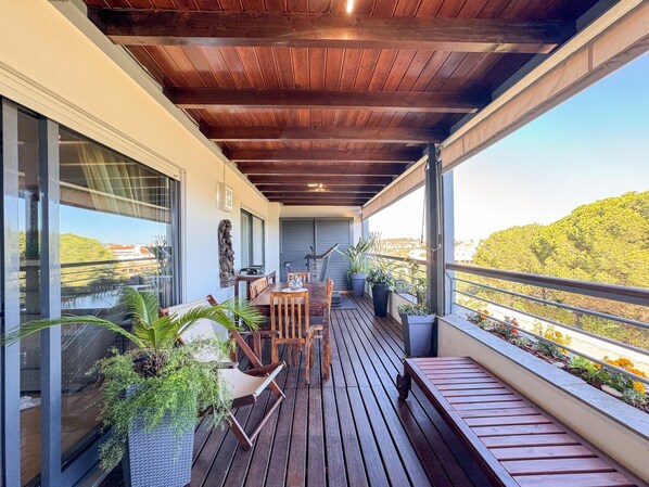 A wooden deck with a table and chairs, potted plants, and a view of a hilly landscape.