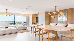 Ein modernes Wohnzimmer mit großem Sofa, Esstisch und Stühlen, einer Küchenzeile und großen Fenstern mit Aussicht.