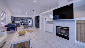 Un amplio salón con chimenea, un televisor de pantalla plana montado encima y una cocina al fondo.