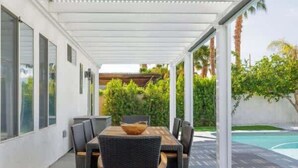 Un patio cubierto con una mesa y sillas de comedor, una piscina al fondo y una pared exterior blanca.