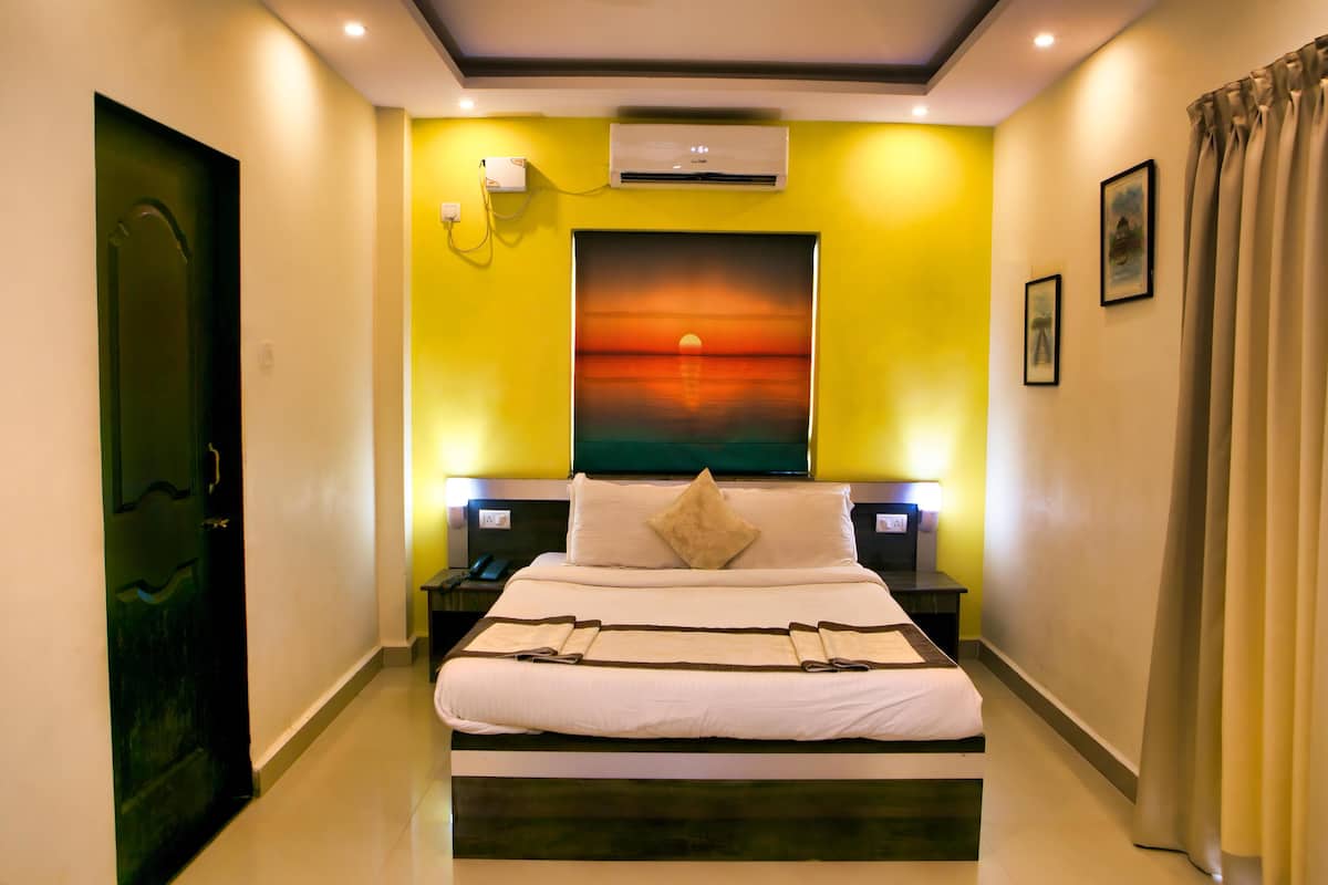 A hotel room with a large bed, a painting of a sunset, an air conditioning unit, and a nightstand with a lamp.
