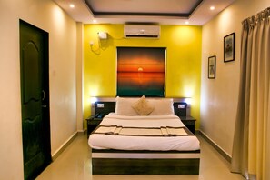 A hotel room with a large bed, a painting of a sunset, an air conditioning unit, and a nightstand with a lamp.