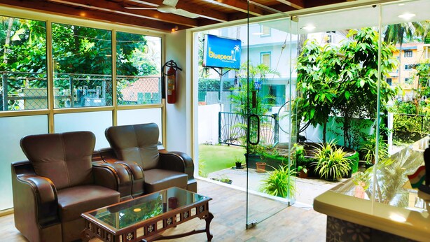 A spacious room with leather sofas, a glass coffee table, and large windows offering a view of a lush garden and a building with a blue sign. - Daksh Elite Candolim Goa (Candolim)