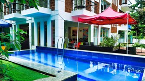 A swimming pool with a red umbrella, surrounded by a building with a white facade and a small garden area. - Daksh Elite Candolim Goa (Candolim)