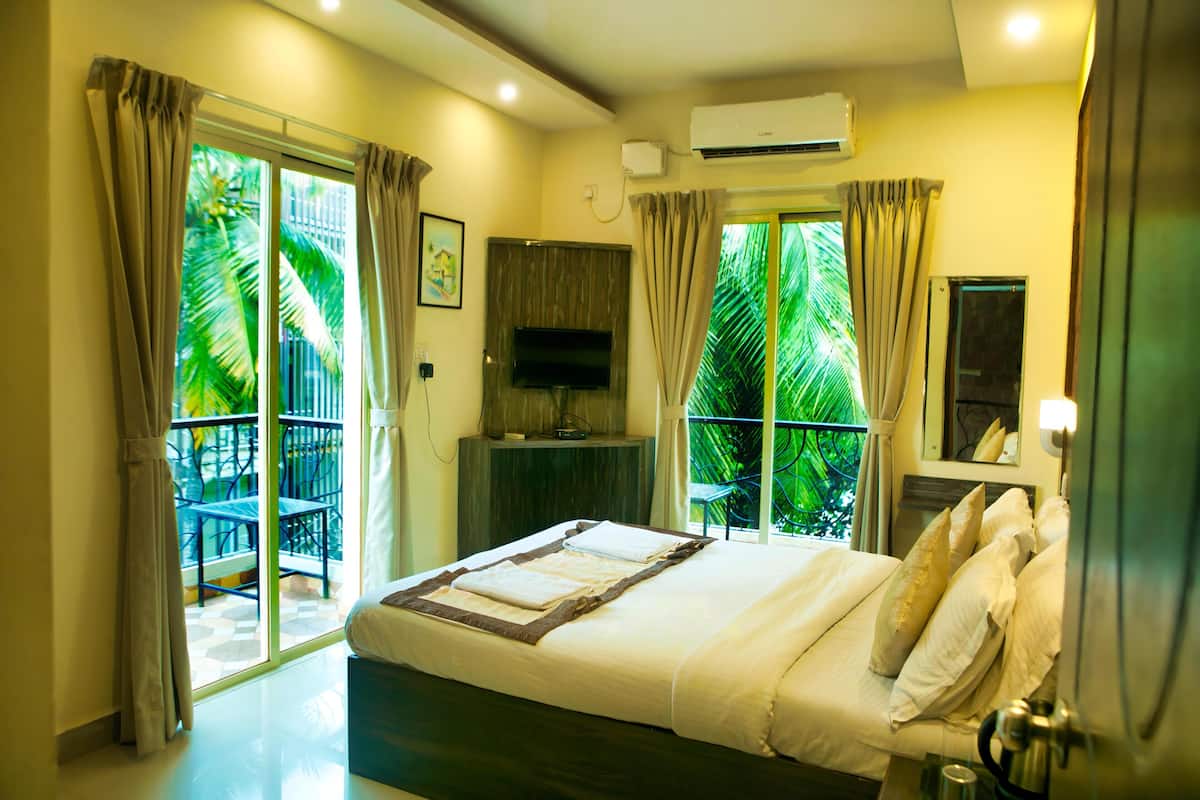 A bedroom with a bed, a TV, a balcony with chairs, and a view of palm trees.