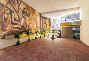 Un vestíbulo amplio con un mural grande, plantas en macetas y una escalera que sube a un nivel superior.