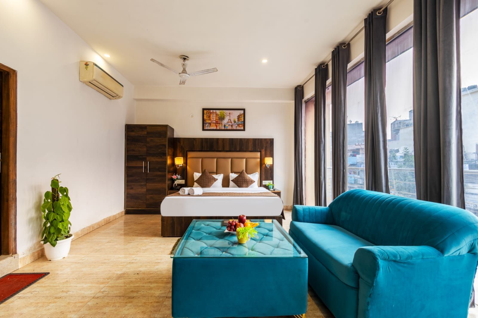 Habitación de hotel con una cama, un sofá azul, una mesa de centro de vidrio con fruta y una planta en maceta, y vistas a la ciudad.