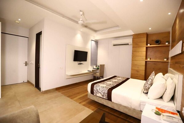 A modern hotel room with a large bed, a flat-screen TV mounted on the wall, a wooden accent wall, and a small desk with a chair. - Hotel Kiara Delhi Airport (New Delhi)
