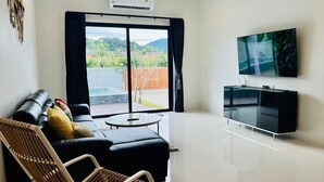 A modern living room with a black sofa, a wicker chair, a flat-screen TV, and a sliding glass door leading to a patio with a hot tub. - Unchan Livliness Pool Villa  (Tambon Ao Nang)