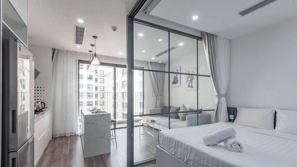 A modern hotel room with a large bed, a small kitchenette, a dining area, and a view of the cityscape. - Coco Apartment Dcapital Luxury (Hanoi)