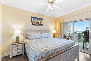 A bedroom with a bed, two bedside tables with lamps, a ceiling fan, a painting, and a balcony with a view. - Crystal Tower 409 (Gulf Shores)