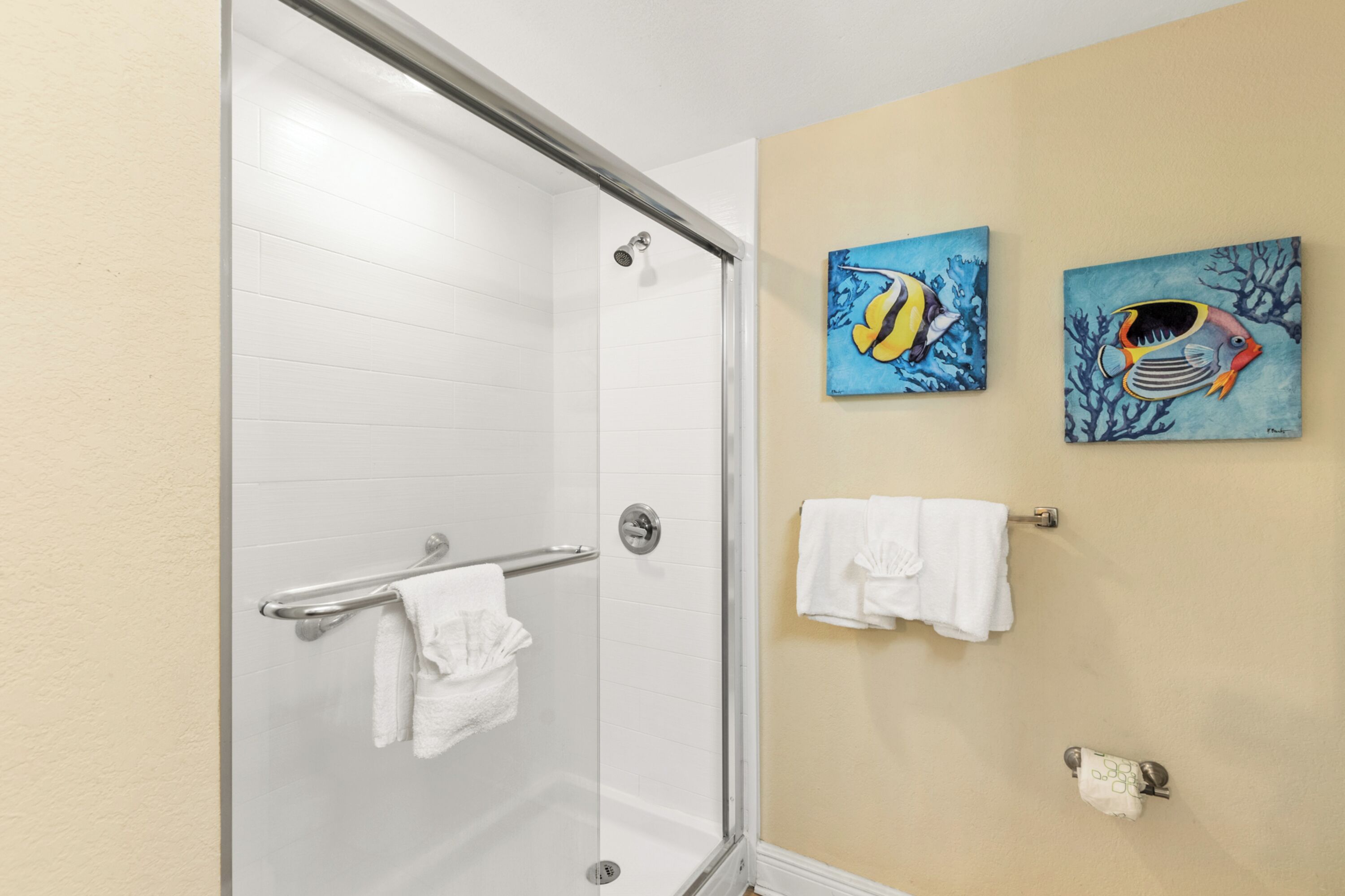 A bathroom with a glass shower door, white tiled walls, and two wall-mounted paintings of fish.