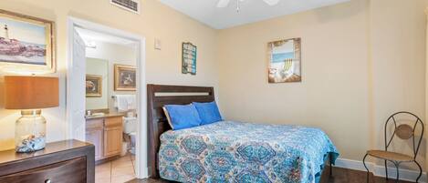 Une chambre à coucher comprenant un lit, une chaise, une table de chevet, une lampe et un ventilateur de plafond.
