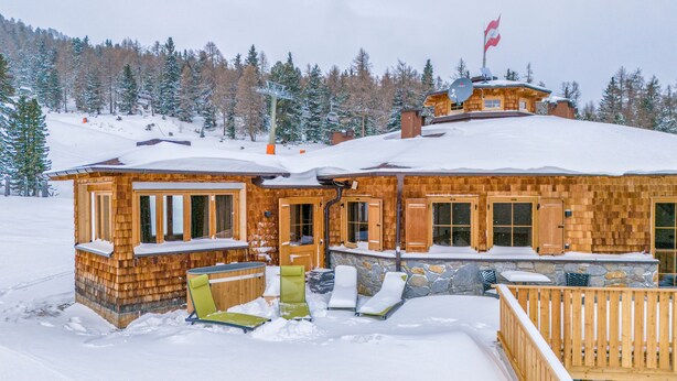 Een houten lodge met een met sneeuw bedekte dak, een skilift op de achtergrond en een besneeerd landschap.