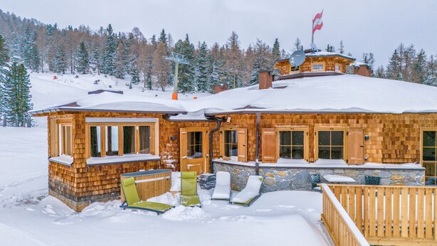 A wooden lodge with a snow-covered roof, a ski lift in the background, and a wooden deck with lounge chairs. - Luxurious apartment with a sauna on the slope (Weißpriach)