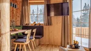 A cozy cabin interior with a wooden dining area, a bench with chairs, a flat-screen TV, and large windows offering a view of trees and snow outside. - Luxurious apartment with a sauna on the slope (Weißpriach)