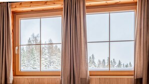 A room with a wooden window and curtains, a bed with a white mattress and a white blanket, and a view of snow-covered trees outside.