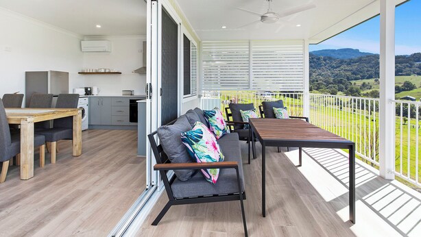 Ein geräumiger Balkon mit Holzboden, einer Couch mit bunten Kissen, einem Esstisch und Blick auf eine grüne Landschaft.