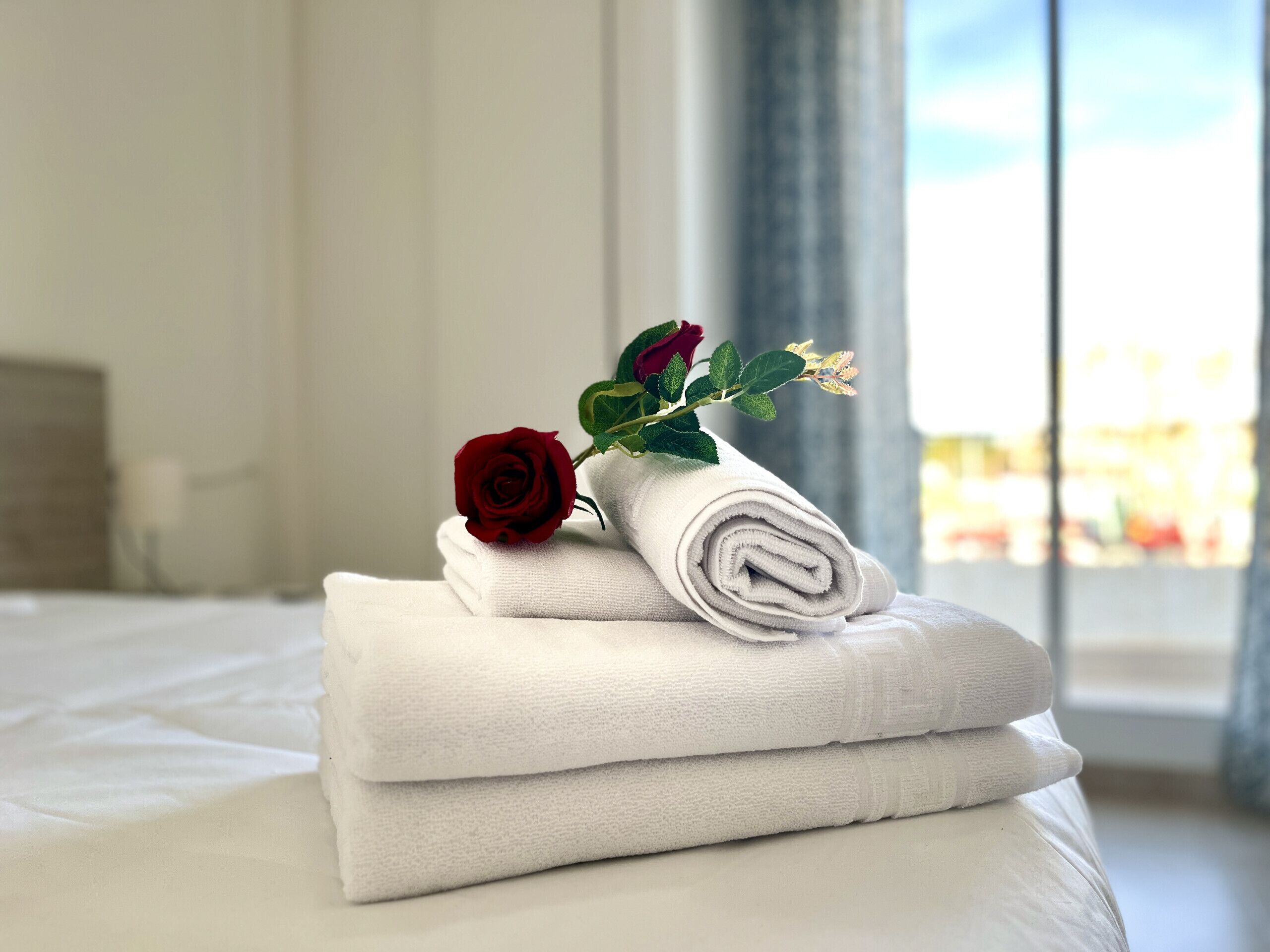 A neatly made bed with folded white towels and a red rose on top.
