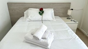 Una cama bien hecha con sábanas blancas, una rosa roja solitaria y toallas dobladas sobre un cojín blanco.