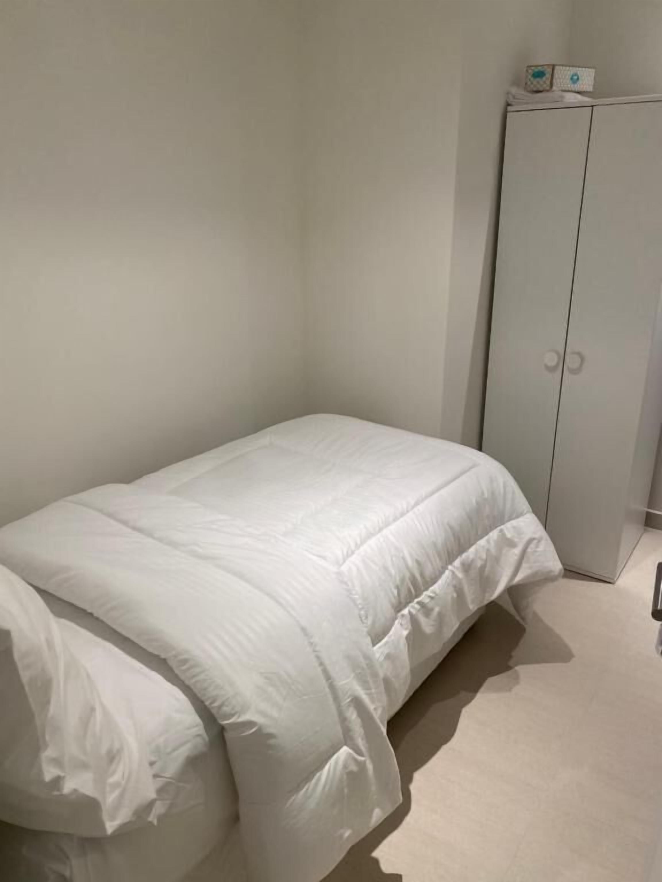 A bedroom with a single bed, a wardrobe, and a small table with items on top.