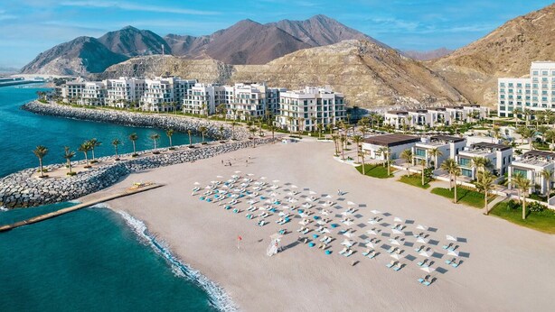 An aerial view of a coastal resort with multiple buildings, a sandy beach with sun loungers, and a rocky breakwater. - Address Residence Fujairah by Bespoke (Sharm)