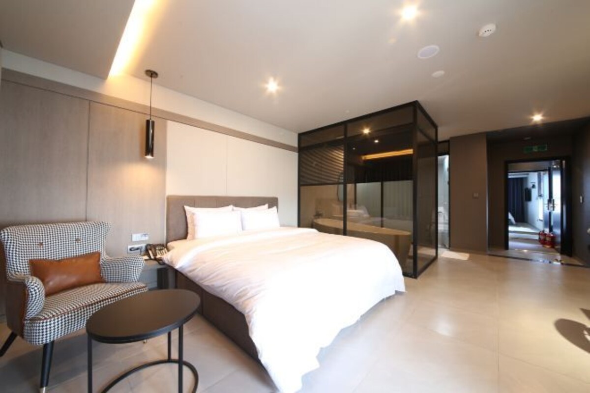 A modern hotel room with a large bed, a checkered armchair, a small table, and a glass partition wall.