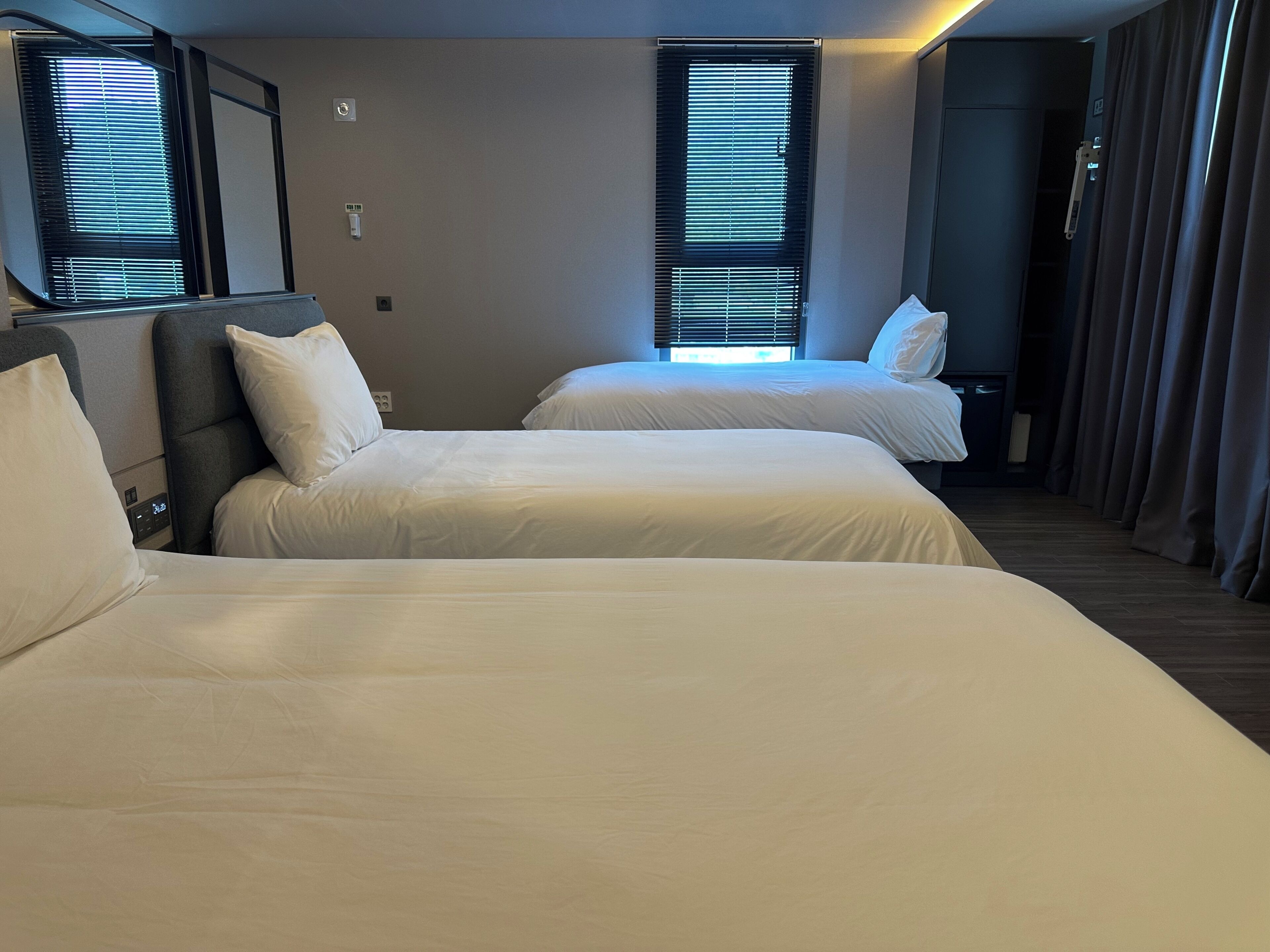 Habitación de hotel con dos camas, cabecera, ventana con persianas y panel de control en la pared, además de una cortina.