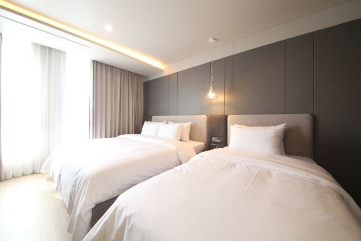 Habitación de hotel con dos camas, un ventanal con cortinas y una lámpara moderna.