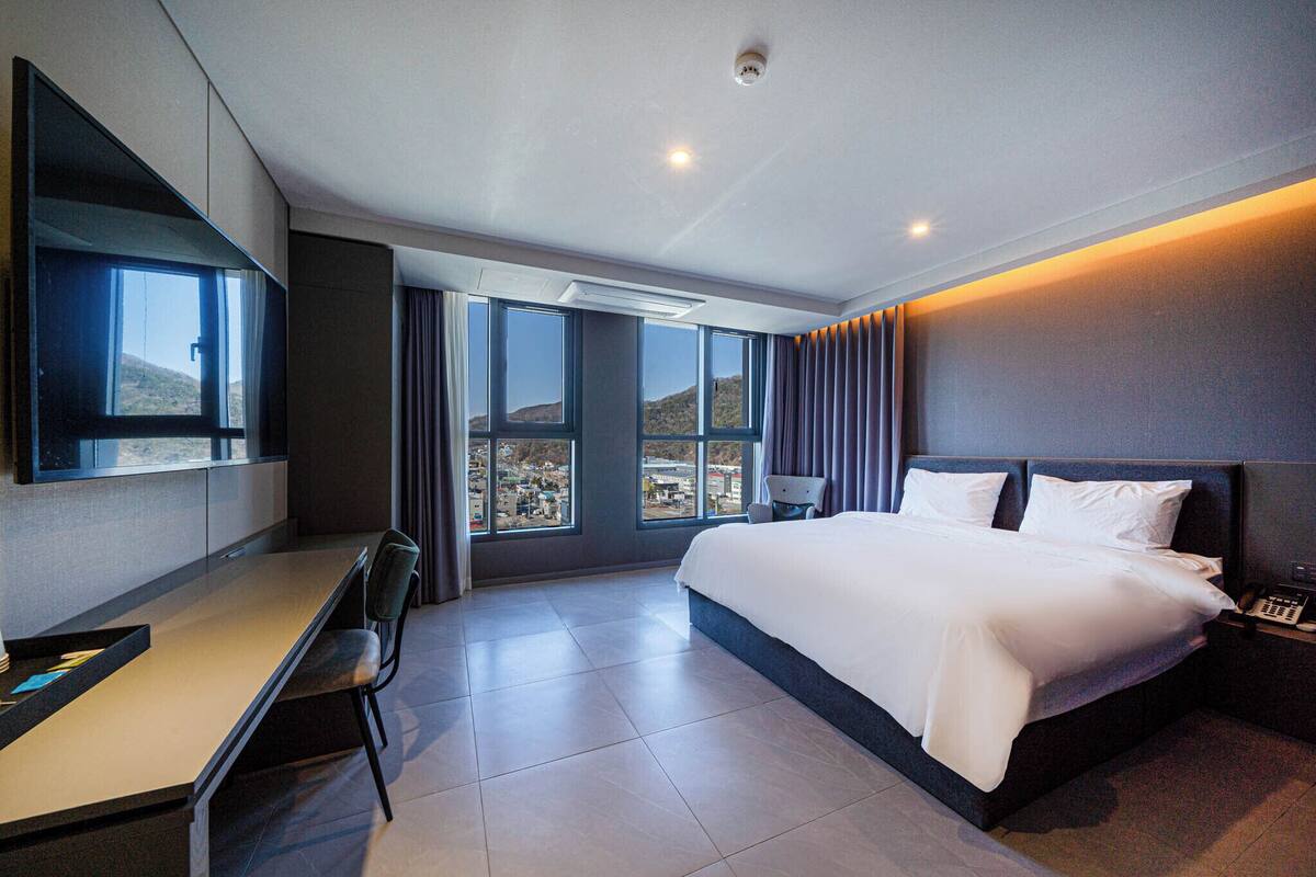 A modern hotel room with a bed, desk, and large windows offering a view of the city.