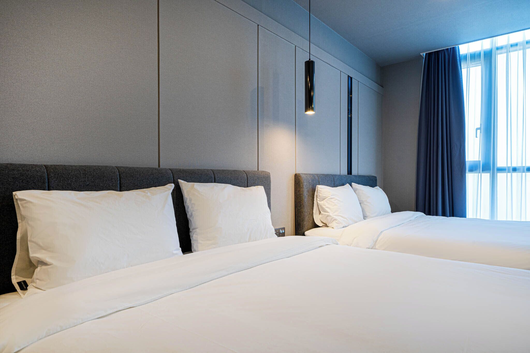 Une chambre d’hôtel avec deux lits, une tête de lit moderne, un luminaire suspendu et une vue sur l’extérieur par une fenêtre agrémentée de rideaux.