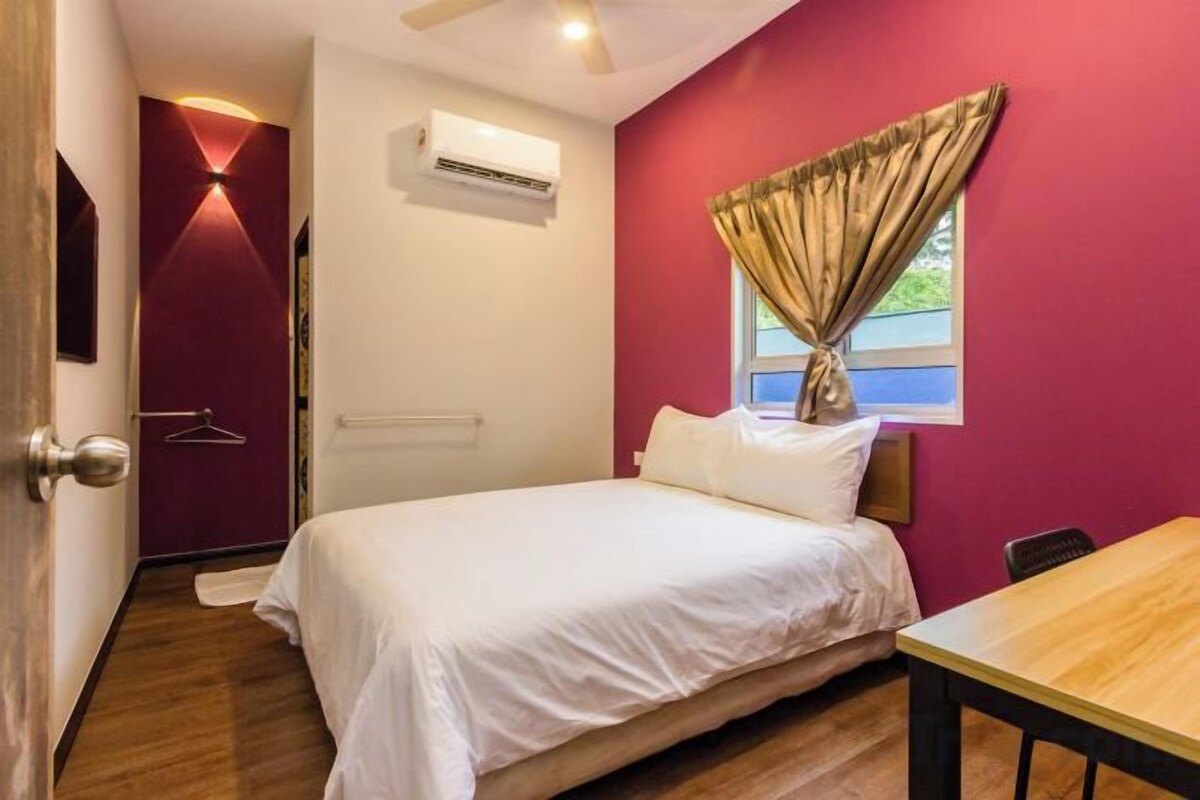 A bedroom with a bed, a window with curtains, a table, and an air conditioning unit.