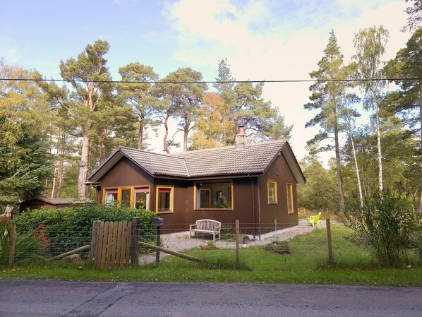 Une maison en bois de couleur brune, avec un toit en tuiles, entourée d’arbres et d’une clôture.