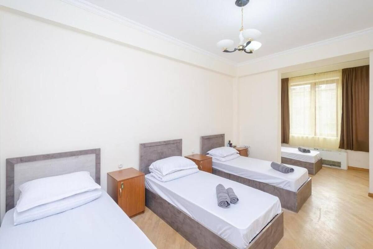 Ein Hotelzimmer mit drei Einzelbetten, Holz-Kopfteilen, einem Nachttisch und einer Nachttruhe, einer Deckenleuchte und einem Fenster mit Vorhängen.