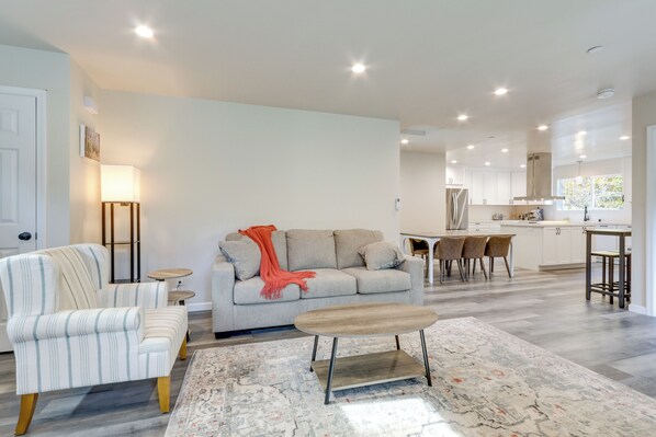 Un salon moderne avec un canapé, un fauteuil et une table basse, un coin repas avec des chaises, et une cuisine en arrière-plan.