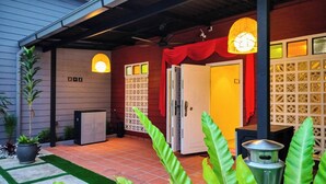 A covered outdoor area with a tiled floor, a small garden with potted plants, and a building with a red door and a yellow door. - Great 5BR Garden Inn by uBook (Butterworth)
