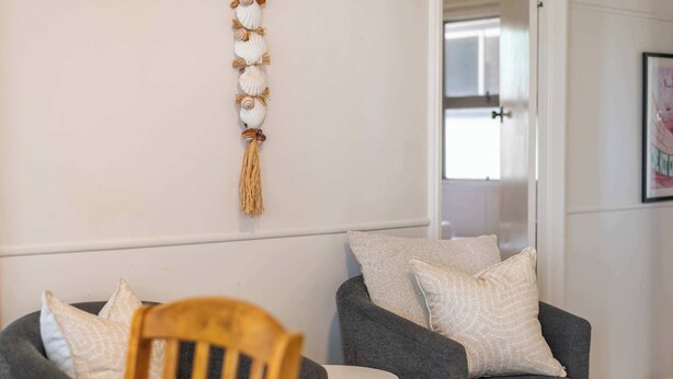 A room with a grey sofa, a wooden chair, a small white table, and a wall-mounted macrame decoration.